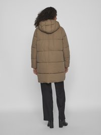 Brun vadderad jacka med huva, med en quiltad design, långa ärmar och knälång fåll, bärs över svarta vida byxor.