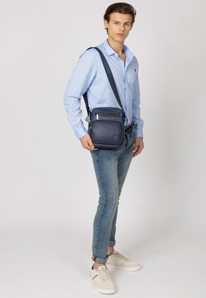 Marinblå læder crossover-taske med justerbar strop, frontlomme med lynlås og præget logo. Modellen bærer en lys blå skjorte og jeans.