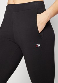 Leggings deportivos negros hechos de una tela elástica, que presentan un bolsillo lateral, una cintura alta y un pequeño logo bordado en colores contrastantes.