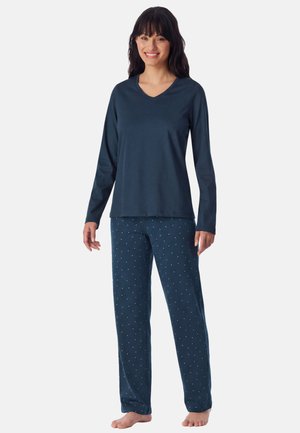 Mujer sonriendo y de pie descalza con camiseta de manga larga azul marino y pantalones de pijama azul marino con pequeños puntos rosas sobre fondo blanco.