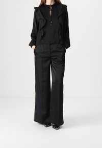 Blouse noire avec des accents à volants et un lien au cou, associée à un pantalon noir taille haute, tous deux en tissu lisse.