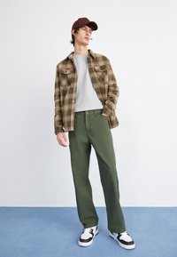 Brown plaid flannel shirt over gray sweatshirt, olive green pants, and black-and-white sneakers. Model stands against a plain background.