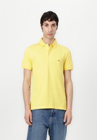 Gelbes Poloshirt mit Kragen und einer Knopfleiste mit drei Knöpfen. Aus Baumwollstoff, mit kurzen Ärmeln, versehen mit einem kleinen gestickten Logo auf der Brust.