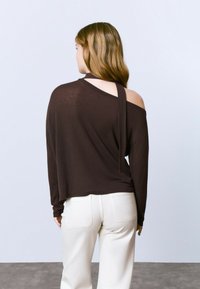 Top marron à manches longues avec un détail dos nu, présentant une encolure large et un tissu doux et léger ; associé à un pantalon blanc taille haute.