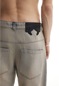 Torso nu d'une personne portant un pantalon en denim gris clair avec deux poches arrière et une pièce en cuir noir sur la ceinture, portant un bracelet en perles.