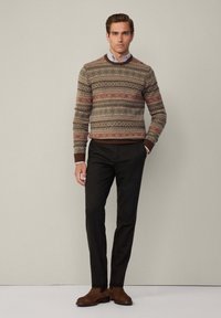 Strickpullover in Erdtönen mit geometrischen Mustern, kombiniert mit schwarzen Hosen und braunen Wildleder-Stiefeletten. Klassisches Hemd mit Kragen sichtbar.