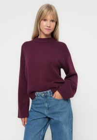Maglione a coste di colore viola scuro, con colletto alto e maniche larghe, abbinato a jeans azzurri a vita alta. Texture morbida.