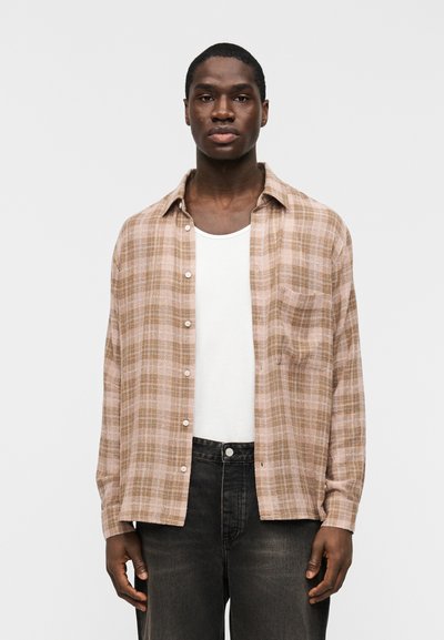 Homme portant une chemise à carreaux beige et marron boutonnée par-dessus un débardeur blanc et un jean noir, debout devant un fond gris clair.