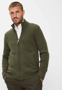 Gebreide groene trui met hoge kraag, ritsluiting en geribde manchetten en zoom. Beschikt over zijzakken en een gemiddelde dikte.