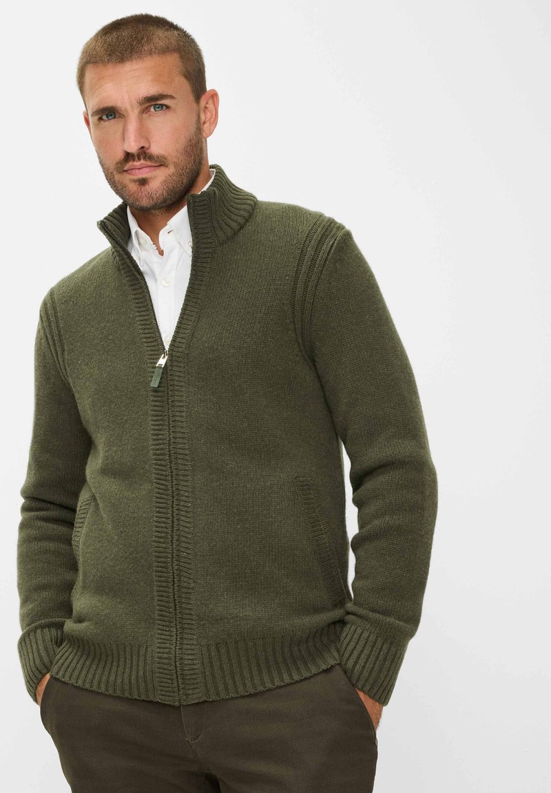 Gebreide groene trui met hoge kraag, ritsluiting en geribde manchetten en zoom. Beschikt over zijzakken en een gemiddelde dikte.