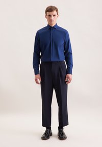 Marineblauwe button-up shirt met een gladde textuur, gecombineerd met geplooide donkere broek en zwarte schoenen, staand op een neutrale achtergrond.