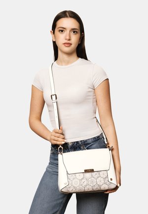 Giovane donna con t-shirt bianca e jeans blu tiene una borsa bianca con pannello frontale decorato e tracolla regolabile.