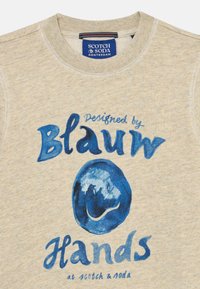 Sudadera ligera de color beige con texto gráfico azul "Diseñado por Blau Hands" y un patrón circular de sonrisas azules. Material de algodón y diseño de cuello redondo.