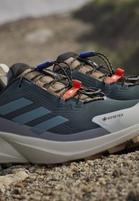 Close-up of dark blue and beige hiking shoes with Gore-Tex fabric and quick lacing system on rocky ground outdoors.