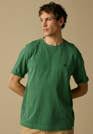 Jeune homme aux cheveux bouclés portant un t-shirt vert à manches courtes et un pantalon beige, regardant vers sa gauche sur un fond beige.