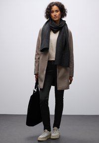 Pitkä tummanharmaa huivi, joka on pukeutunut vaalean kermaiseen villapaitaan, yhdistettynä beigeen takkiin, mustiin farkkuihin ja beigeihin lenkkareihin. Musta laukku kädessä.