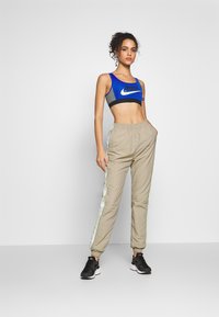 Sportstopp i blått med svarte og grå detaljer, kombinert med beige joggebukser med en hvit og grønn sidestripe. Svart treningssko.