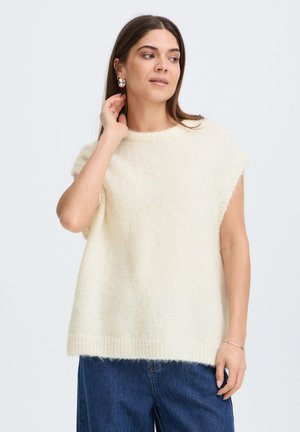 Femme portant un pull sans manches, duveteux écru et un jean bleu, touchant son oreille et regardant sur le côté devant un fond uni.
