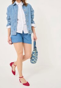 Femme portant une veste en jean et un short, une chemise blanche boutonnée, des chaussures rouges à bride à la cheville, et portant un sac à main bleu texturé.