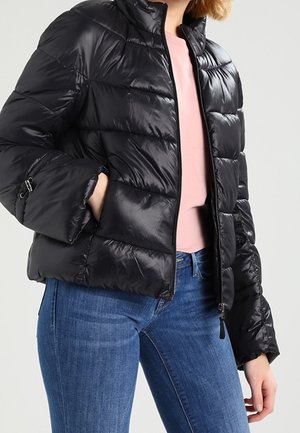 Femme portant une doudoune noire brillante par-dessus un haut rose et un jean bleu, avec une main dans la poche de la veste.