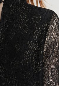 Blusa nera in pizzo con delicati motivi floreali, caratterizzata da un alto colletto e maniche trasparenti, arricchita da sottili fili d'oro.