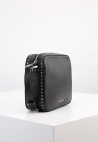 Svart läder crossbody-väska med strukturerad yta, fyrkantig form, silverfärgade detaljer, dragkedjestängning och öljettdekoration längs sidorna.