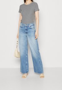 Grå, slim-passande t-shirt och ljusblå, vida jeans med en urtvättad look. Modellen håller en beige handväska och bär beige blockklack-sandaler.