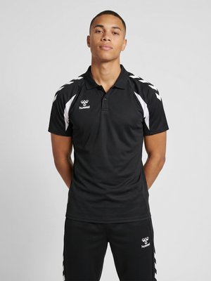 Jonge man staat met handen achter de rug, draagt een zwarte Hummel sportpolo en bijpassende zwarte broek met witte chevron-details.