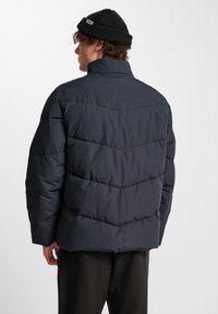 Puffjacka i mörk marinblå, med hög krage och chevron-quiltat mönster. Tyget ser slätt och lättviktigt ut. Svart mössa bärs.