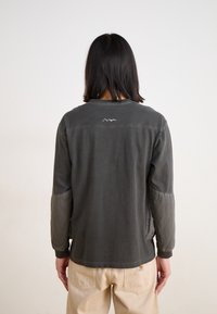 Gray long-sleeve shirt with quilted gray sleeves and a round neckline. Features a small logo on the upper back. Worn with beige pants.