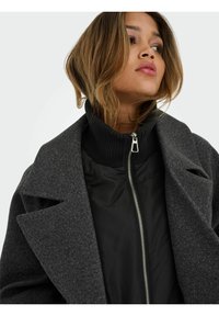 Szary płaszcz z mieszanki wełny z szerokim kołnierzem na czarnej kurtce na zamek. Charakteryzuje się prążkowanym czarnym golfem i srebrnym detalem zamka.