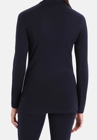 Långärmad topp i mörk marinblått tyg med en figursydd design, slät textur och hög hals. Har sprund på sidorna vid fållen.