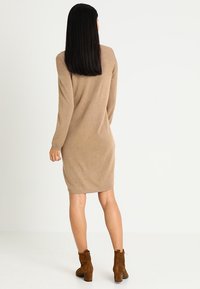Robe en maille à manches longues couleur beige clair, longueur jusqu'aux genoux. Texture douce, coupe droite, détail au dos visible. Portée avec des bottines marron.