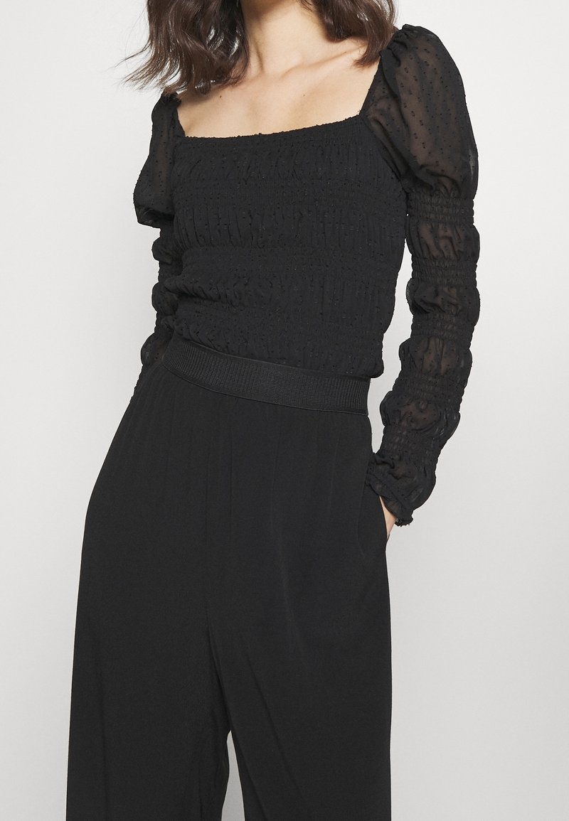 Femme portant un top froncé noir avec des manches longues bouffantes transparentes et un pantalon large noir taille haute avec les mains dans les poches.