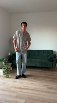 Košeľa s krátkym rukávom a gombíkmi, s svetlými a tmavými pruhmi, v kombinácii s svetlo modrými džínsami a čiernymi topánkami; stojí v miestnosti s zeleným gaučom.