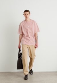 Light pink oversized t-shirt with a small white logo, beige trousers, carrying a black tote bag, worn with black shoes.