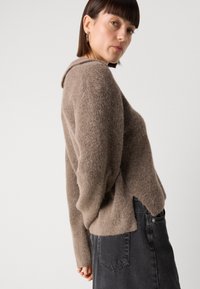Pull en maille marron doux avec une coupe décontractée et un col montant. Doté de poignets côtelés et d'un ourlet asymétrique. Associé à une jupe en denim noire.