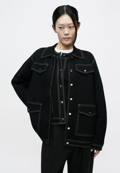 Mustast kootud jakk kontrastsete valgete õmblustega. Omadusteks kaks rinnataskut, nööbitav eesmine osa ja lõdvestunud istuvus.