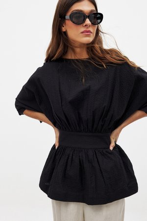 Čierna textúrovaná blúzka so zberaným pásom a širokými rukávmi, skombinovaná s svetlými nohavicami a prehnanými čiernymi slnečnými okuliarmi.