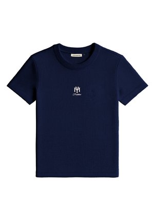 Young man wearing a beige cap with "Champagne Courtside Prohibited" and a black short-sleeve shirt with a white emblem, standing against white background.