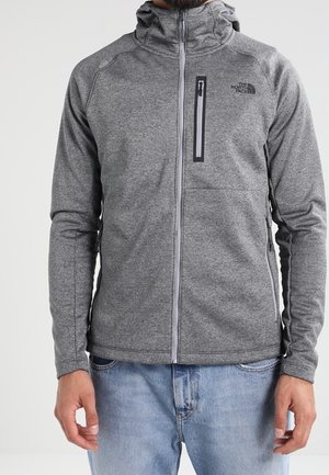 Mężczyzna w szarej bluzie z kapturem i zamkiem błyskawicznym, z kieszenią na piersi i logo The North Face, w połączeniu z jasnoniebieskimi jeansami.