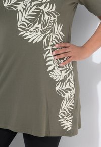 Hand ruht auf der Hüfte, trägt eine olivgrüne Tunika mit weißem Blattmuster und schwarze Leggings, vor einer hellgrauen Wand.