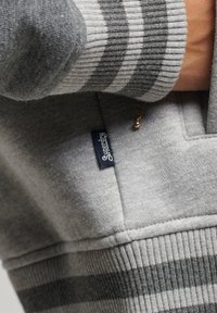 Sweat-shirt gris avec poignets rayés, doté d'une petite étiquette logo "Superdry" bleue. Tissu doux et texturé avec une coupe décontractée.