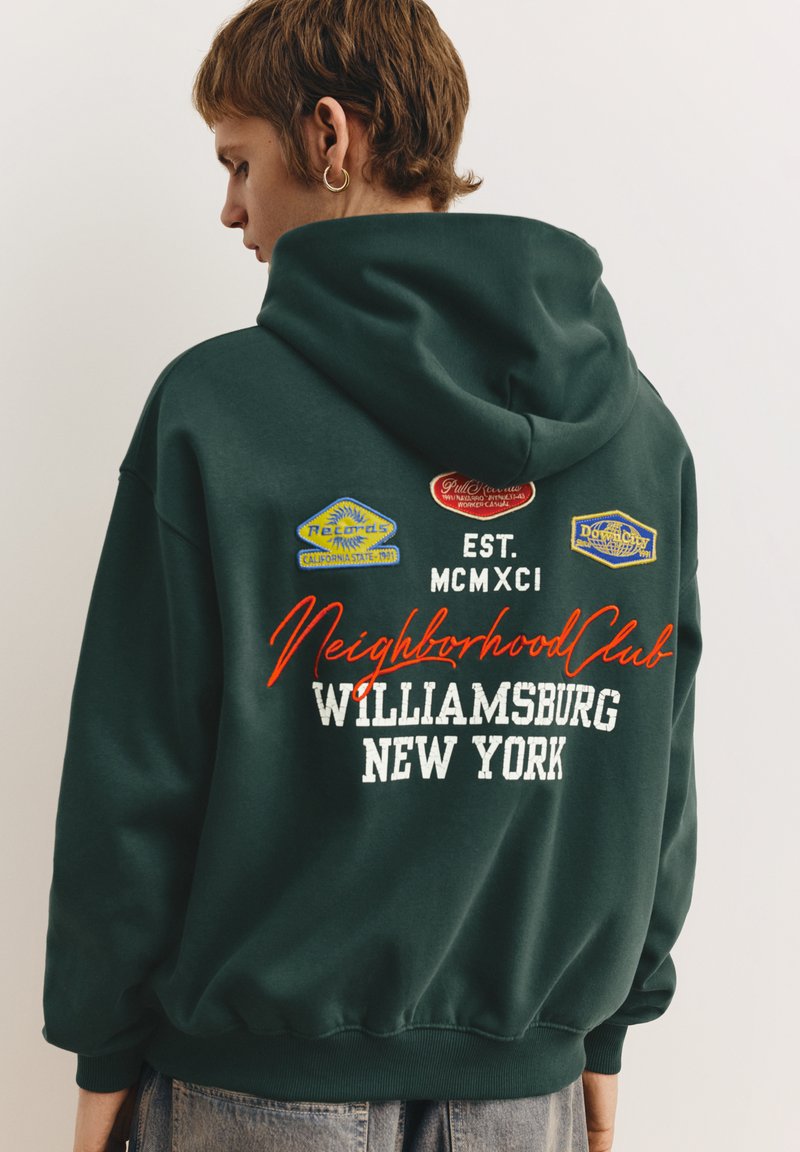 Mørkegrøn hættetrøje med broderede mærker og hvid tekst der siger "Neighborhood Club Williamsburg New York." Fremstillet af bomuldsblanding.