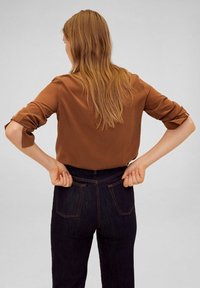 Blouse marron à manches retroussées associée à un jean foncé en denim. Les caractéristiques notables comprennent des coutures contrastées et des poches à l'arrière du jean.