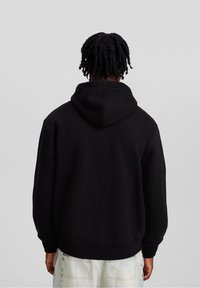 Person med mörkt, vridet hår som bär en svart huvtröja och ljusblå jeans, står vänd bort mot en enkel bakgrund.