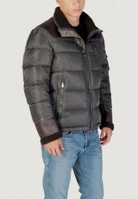 Uomo che indossa un giubbotto imbottito nero con dettagli in pelle marrone e shearling, abbinato a jeans denim blu chiaro, in piedi davanti a uno sfondo bianco.