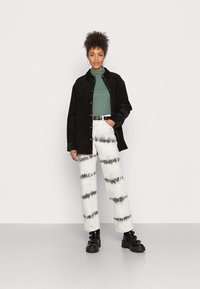 Camisa de veludo cotelê preta sobre um pulôver de gola alta verde, combinada com calças brancas de cintura alta com riscas de tie-dye em preto e botas chunky pretas.