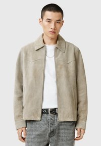 Mockasinfärgad jacka i ljusbeige med spetsig krage, dragkedjestängning och bröstfickor. Tår över en vit skjorta och grå jeans. Slät textur.