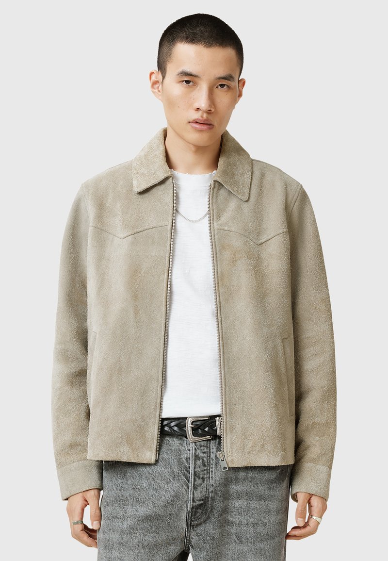 Suède jasje in lichtbeige met een puntige kraag, ritssluiting en borstzakken. Gemaakt over een wit shirt en grijze jeans. Vriendelijke textuur.